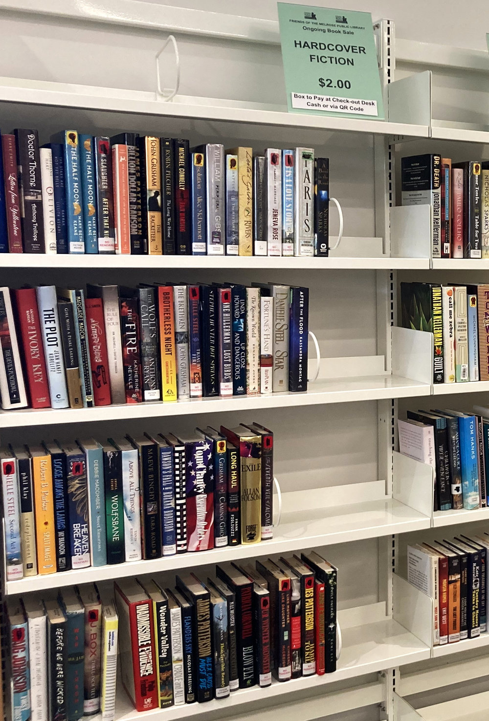 Library shelves of hardcover novels under a sign: Friends of the Melrose Public Library Ongoing Book Sale, Hardcover Fiction, $2.00